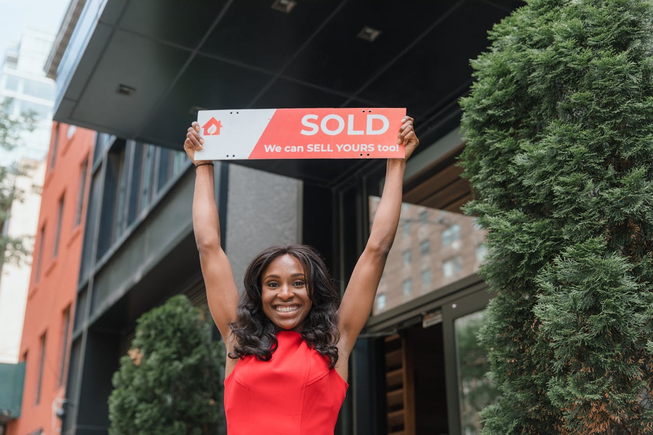 Smiling woman holding a 'Sold' sign outside a building, celebrating a successful sale.